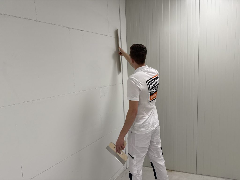 Stukadoor werkt met spaan en plakspaan een lange wand af in een lege ruimte
