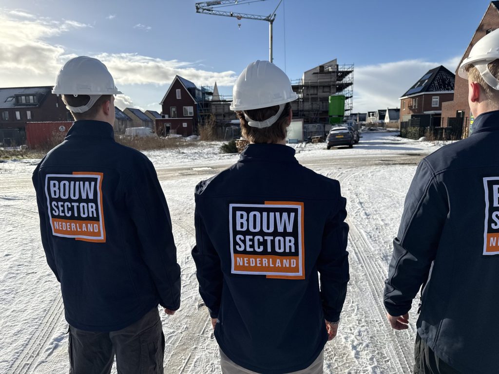 Drie vakmensen van Bouwsector Nederland in blauwe jassen bij een bouwplaats met een hijskraan